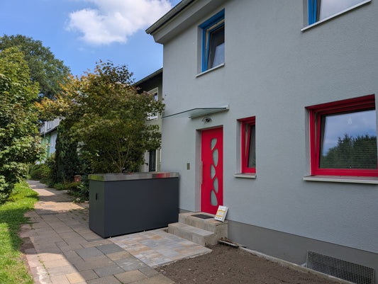 Fahrradgarage neben dem Eingang eines Hauses mit roter Tür und Pflanzen im Vorgarten.