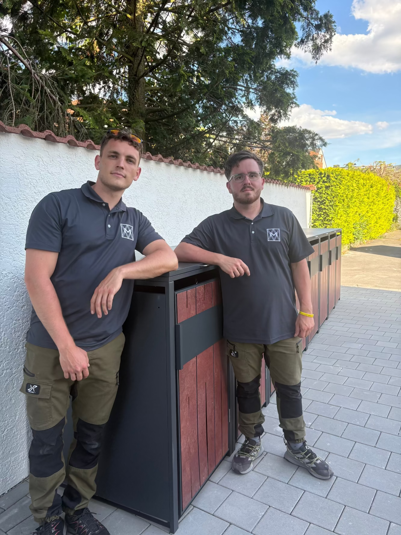 Zwei professionelle Handwerker des Montage-Services stehen stolz neben einer frisch aufgebauten, anthrazitfarbenen Mülltonnenbox mit Holzdekor-Fronten im Außenbereich.