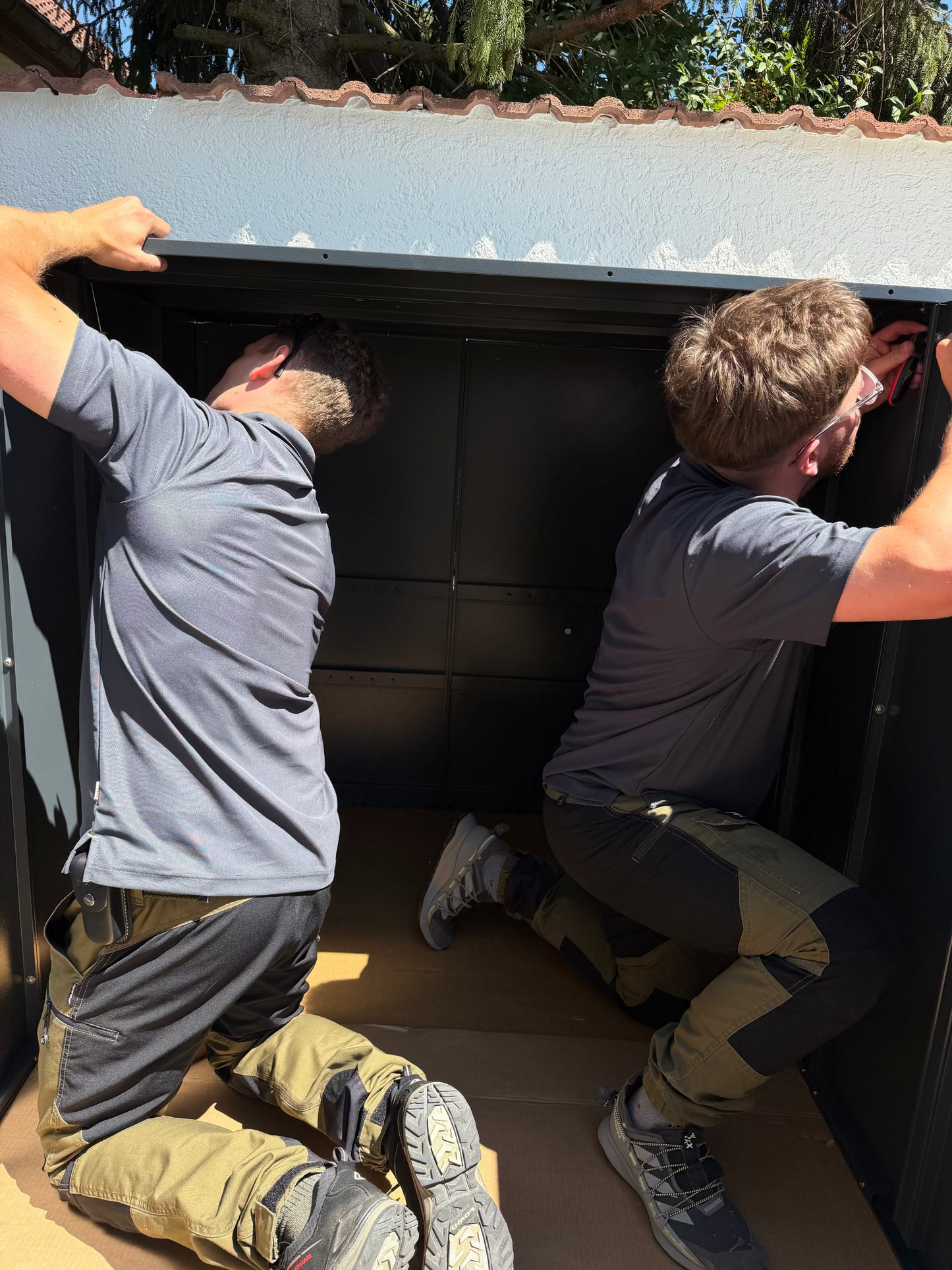 Professioneller Montage-Service für Metallwaren in Deutschland: Zwei Handwerker montieren eine anthrazitfarbene Metall-Gerätebox im Außenbereich.