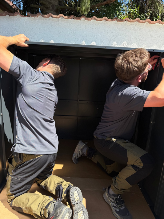 Professioneller Montage-Service für Metallwaren in Deutschland: Zwei Handwerker montieren eine anthrazitfarbene Metall-Gerätebox im Außenbereich.