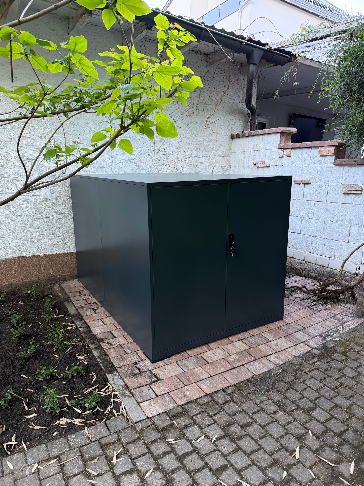 Moderne Metall-Fahrradgarage Container in Anthrazit mit geschlossenen Flügeltüren, aufgebaut auf einer gepflasterten Fläche im Garten.