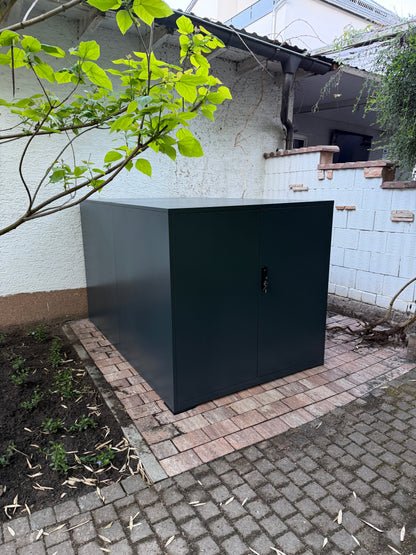 Moderne Metall-Fahrradgarage Container in Anthrazit mit geschlossenen Flügeltüren, aufgebaut auf einer gepflasterten Fläche im Garten.