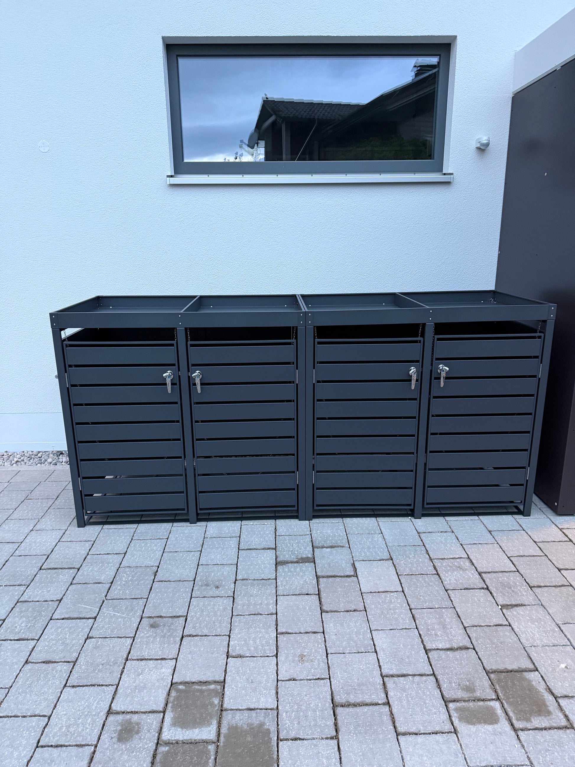 Metallverkleidung für 4 Mülltonnen in Anthrazit mit horizontalen Lamellen, Griffen mit Schlüsseln und integrierten Pflanzwannen, aufgestellt auf Pflastersteinen vor einer weißen Hauswand.
